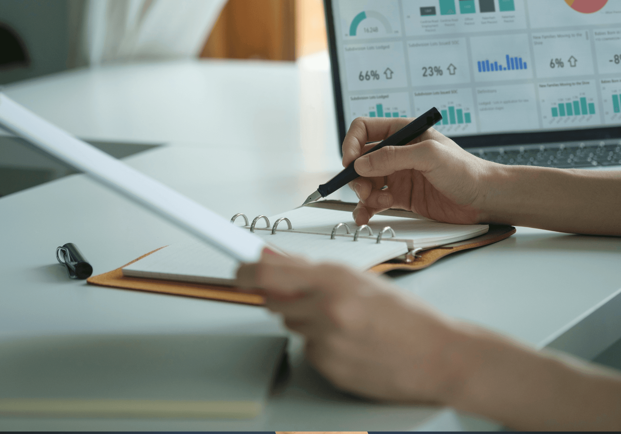 Ter illustratie van administratieve lasten een hand die een pen vasthoudt en in een boekje schrijft met een computerscherm met tabellen op de achtergrond