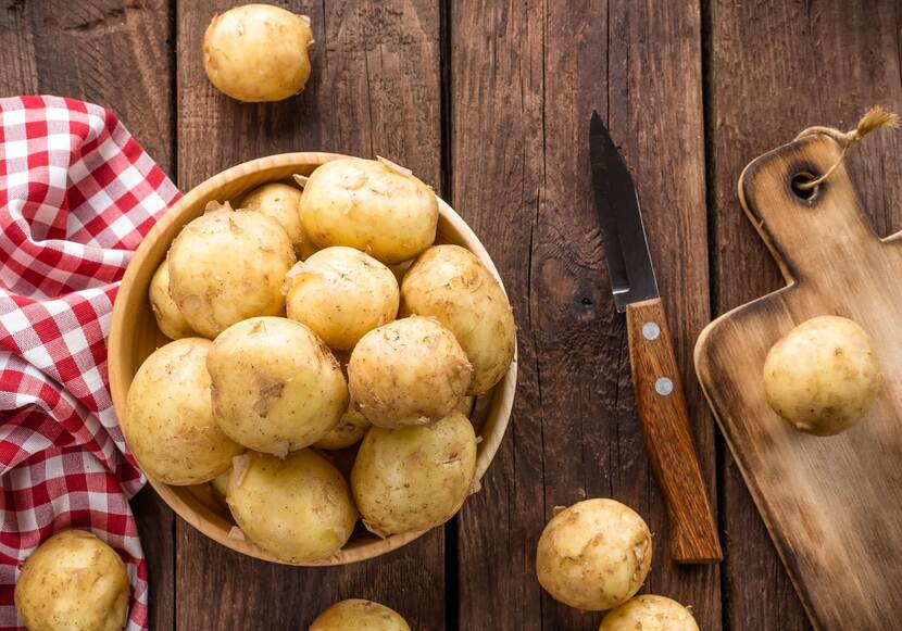 Houten tafel met daarom een links een rood-wit geblokte handdoek, in het midden een schaal met aardappelen en rechts een mes met snijplank en een aardappel erop