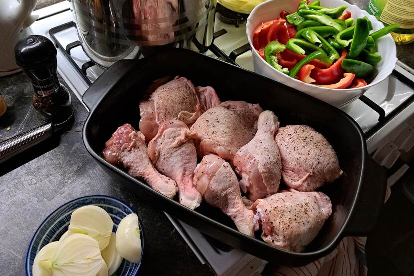 Raw, seasoned chicken legs are placed in a ceramic bowl, ready to be cooked, placed on top of an oven. There are chopped onions and bell peppers in separate bowls.