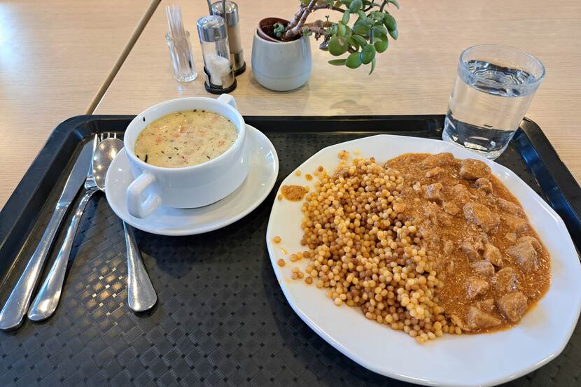 A cafeteria lunch can be seen. The cuisine is Hungarian. There is a small bowl of chicken soup and a plate with Hungarian goulash, on a tray. There are also utensils, a glass of water, there is salt and pepper on the table and a decorative plant.