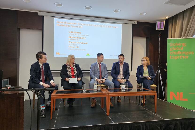 A panel of four experts sit on a raised stage, facing the audience. One expert is talking and explaining something. There is a slide behind the presenters listing their names. There is a Dutch roll-up banner on their left.