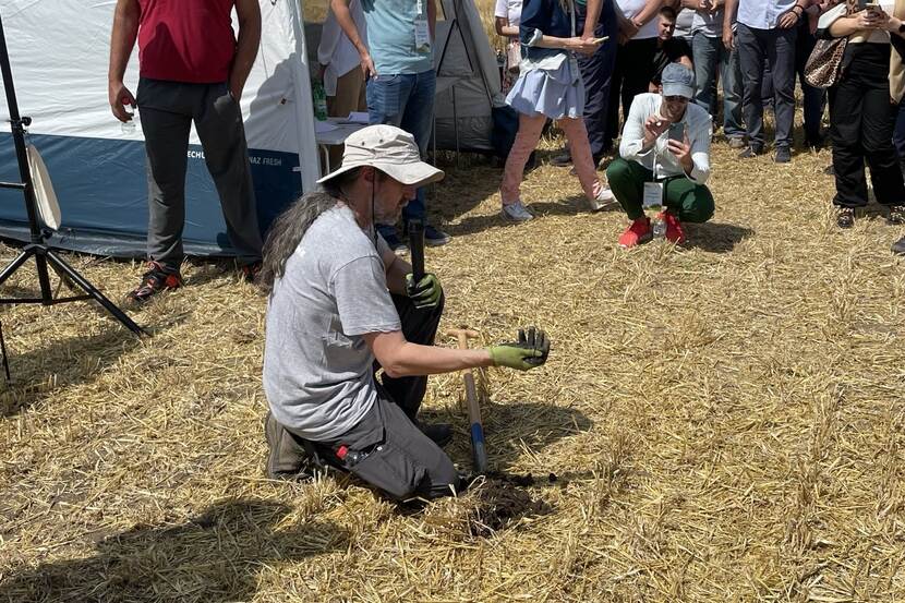 A man wearing a wide-brimmed cloth hat kneels on a field, picking up dirt from the ground and explaining things to an audience.